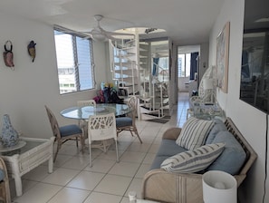 Un vaste espace de vie comprenant une table ronde en verre, des meubles en osier, un canapé agrémenté de coussins à rayures et un escalier en colimaçon.