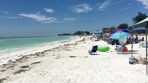 Une plage de sable blanc, avec une eau turquoise et limpide, et quelques parasols qui offrent de l’ombre.