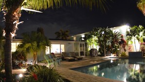 Un espace piscine aménagé pour la nuit, avec des chaises longues, une piscine et un bâtiment bien éclairé en arrière-plan.