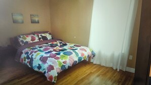 A bedroom with a wooden floor, a bed with a colorful floral bedspread, and a white curtain on the window.
