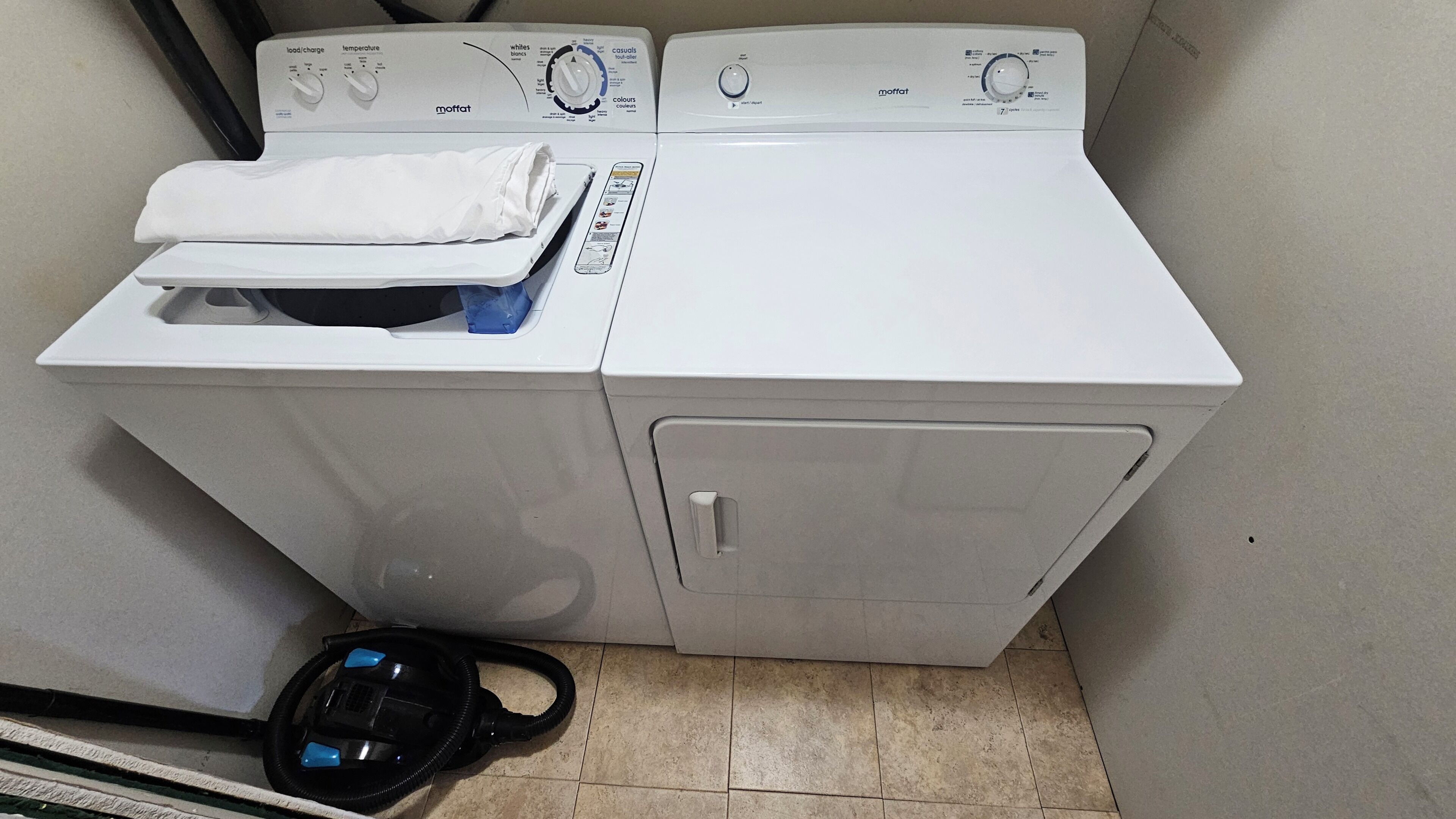 A front-loading white washing machine with a control panel, a white towel on top, and a black vacuum cleaner beside it.