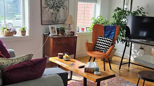 A cozy living room with a wooden coffee table, a comfortable armchair with a patterned pillow, a television, and a window with a view of a car and a brick wall.