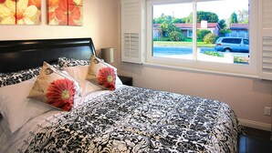 Een slaapkamer met een groot bed, een raam met zonwering en twee bloemschilderijen aan de muur.