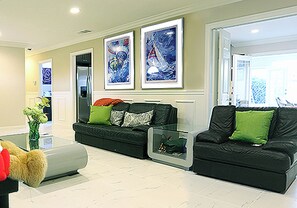 Ein großzügiges Wohnzimmer mit einem großen Teddybären, einer Couch und einem Couchtisch. An den Wänden befinden sich gerahmte Kunstwerke und eine Tür zu einem weiteren Raum.