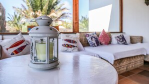 A white table with a lantern containing a candle, a white sofa with colorful pillows, and a view of palm trees through large windows. - VILLA VOLCANO- Rural peaceful Gem under the Volcano  (LAJARES)