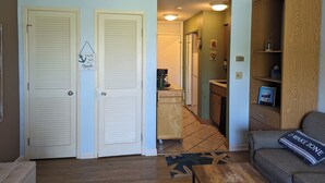 A living room with a sofa, a coffee table, and a bookshelf. There are two doors, one leading to a hallway and the other to a kitchen. - Make memories on Grand Traverse East Bay. (Williamsburg)