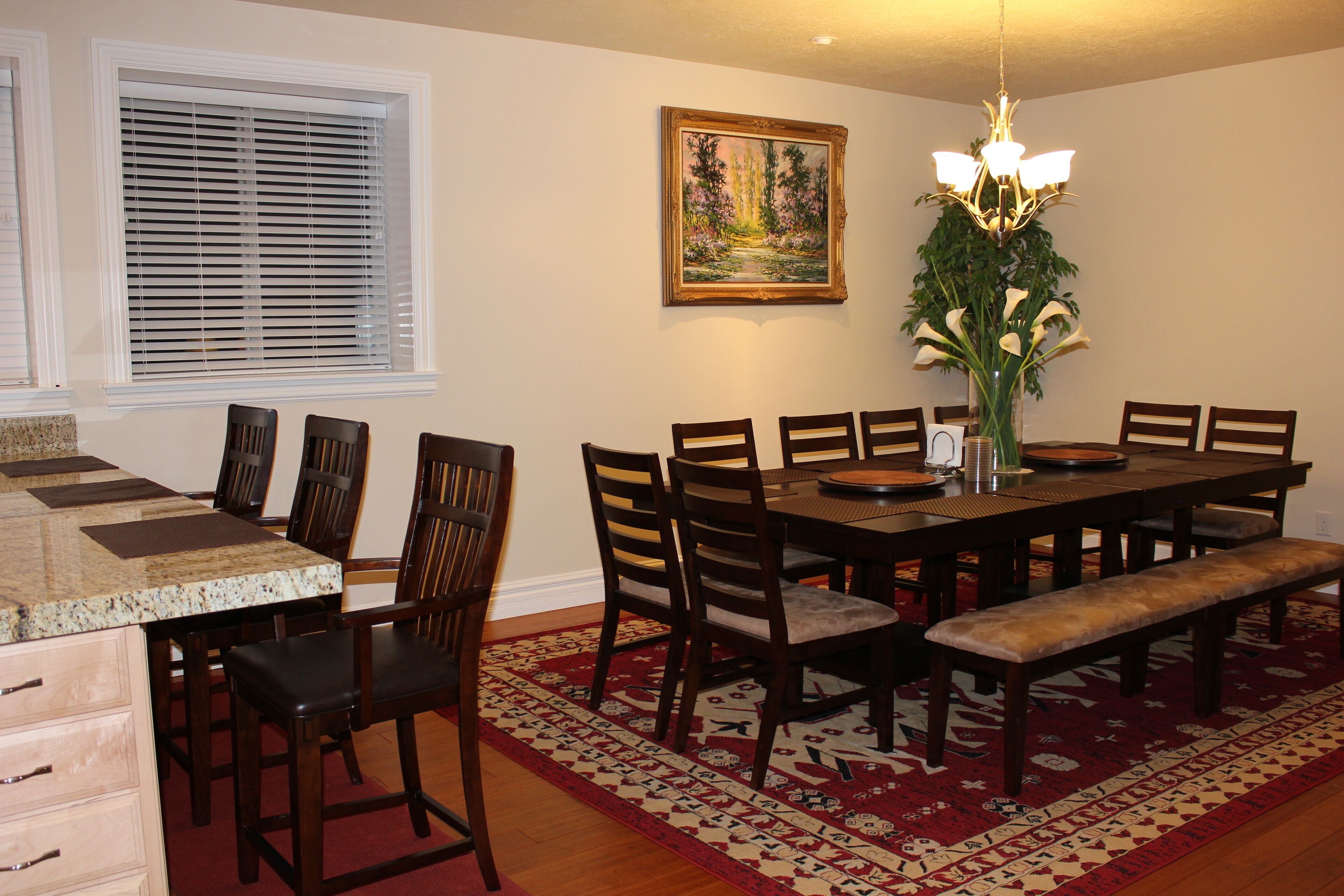 Un coin repas comprenant une table en bois, des chaises, un banc, un tapis, un tableau et un lustre.