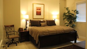 Un dormitorio con una cama, dos mesitas de noche, una silla, un ventilador de techo, un cuadro en la pared y una ventana con persianas.