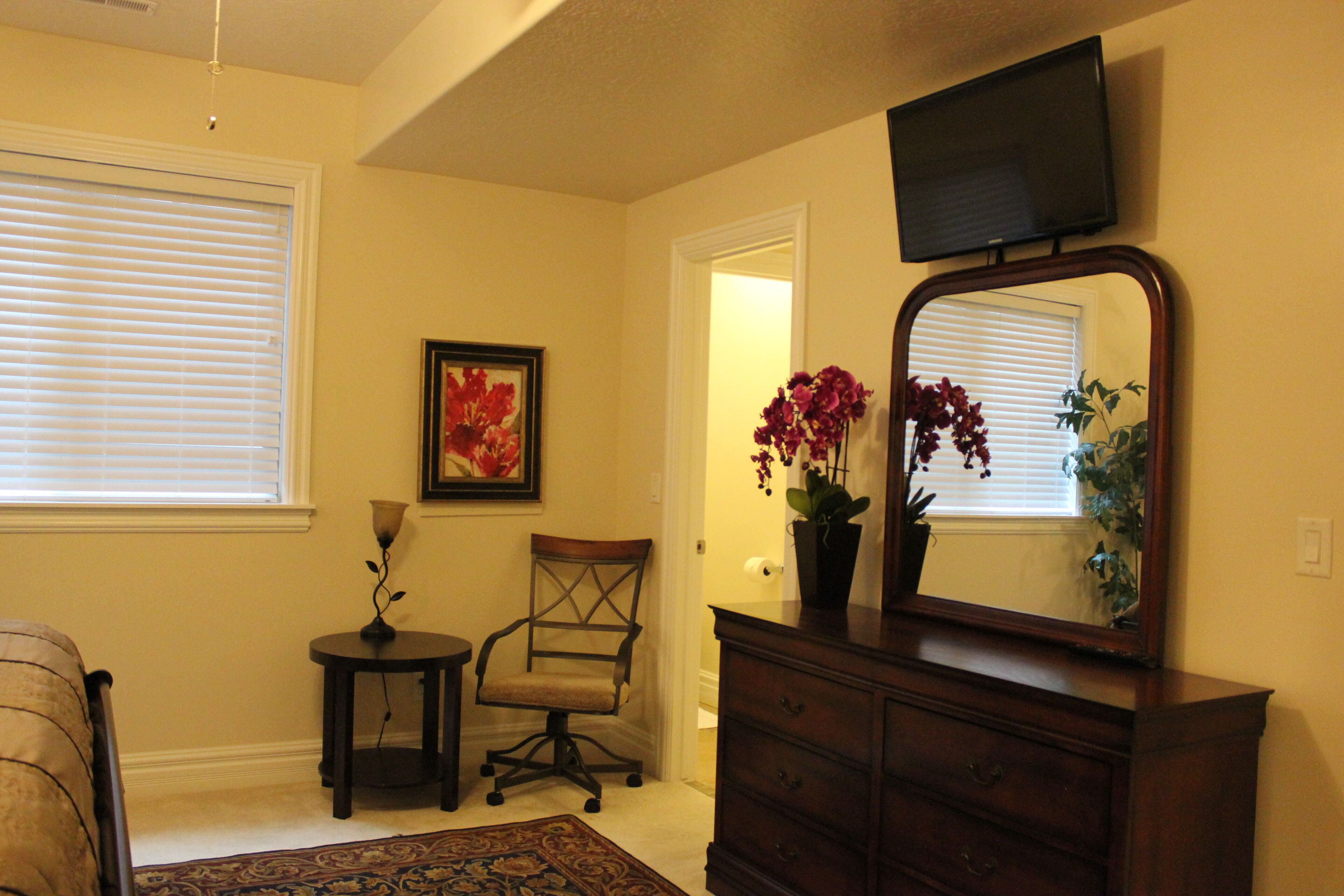 A bedroom with a bed, a dresser, a chair, a small table, a mirror, and a TV mounted on the wall.