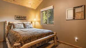 Un dormitorio con cabecera de cama de madera, colcha estampada, mesita de noche, lámpara, ventana con vistas a árboles y espejo adosado a la pared.