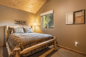 A bedroom with a wooden bed frame, a patterned bedspread, a nightstand, a lamp, a window with a view of trees, and a wall-mounted mirror.