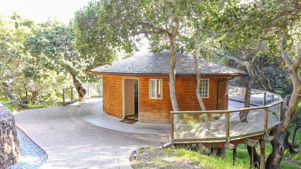 A circular wooden cabin with a glass balcony, surrounded by trees and a paved path.