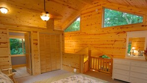 A wooden cabin bedroom with a bed, dresser, and a window overlooking greenery.