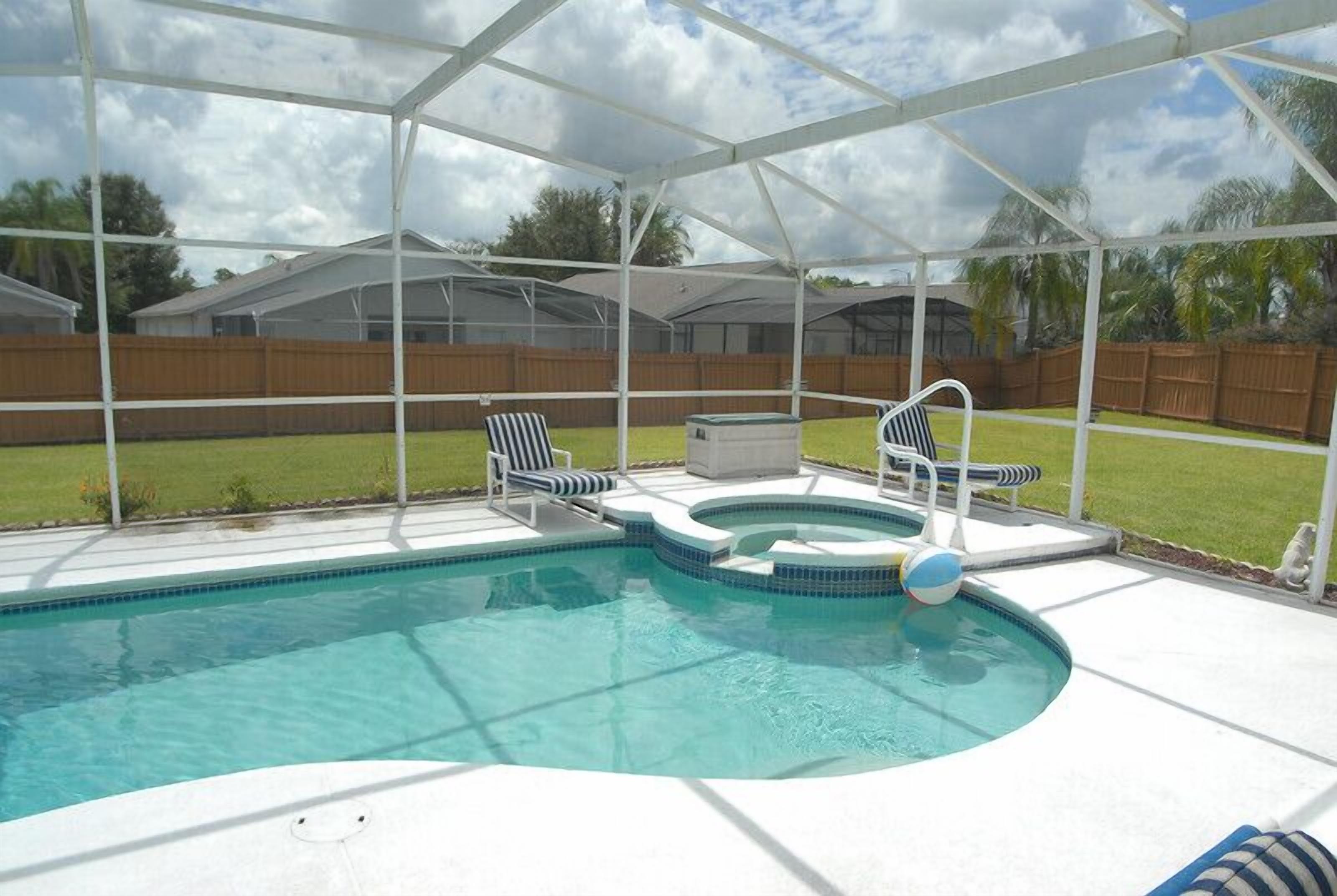 A covered outdoor swimming pool with a hot tub, lounge chairs, and a wooden fence in the background.