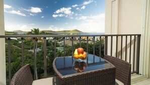 A balcony with a glass table and wicker chairs, overlooking a scenic landscape with hills and a body of water. - Paradise @ Sapphire Village Studio- H-S WIFI -No Excess Owner Fees #307 (St Thomas)