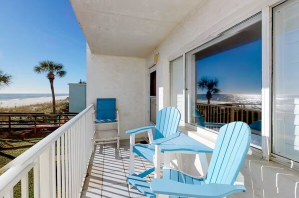 Ein Balkon mit zwei blauen Stühlen und einem kleinen Tisch, mit Blick auf einen Strand mit Palmen und einem klaren Himmel.