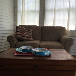 A brown sofa with a patterned pillow, two blue bowls on a wooden table, and sheer curtains in a room with white walls.