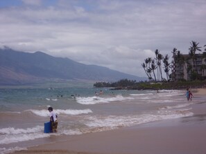 A beach with people swimming and walking along the shore, palm trees, and mountains in the background. - SALE! -- Best Location  &  Closest to the Beach - One Bedroom (kihei)