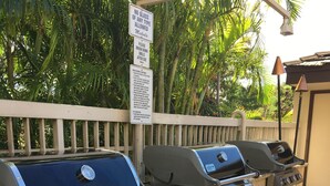 A barbecue grill setup with multiple burners and a built-in oven, located outdoors with a wooden fence and lush greenery in the background. - SALE! -- Best Location  &  Closest to the Beach - One Bedroom (kihei)