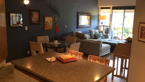 A modern living room with a dining area, featuring a glass-top table, comfortable seating, and artwork on the walls. - SALE! -- Best Location  &  Closest to the Beach - One Bedroom (kihei)