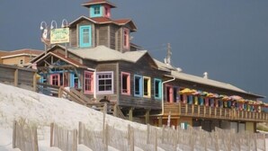 A multi-story building with a wooden facade and colorful accents, featuring a prominent sign and a balcony with umbrellas. - Upscale Home, Pool, Hot Tub, EV Charger, Pets OK, 10% Off Golf Cart Rentals (Miramar Beach)