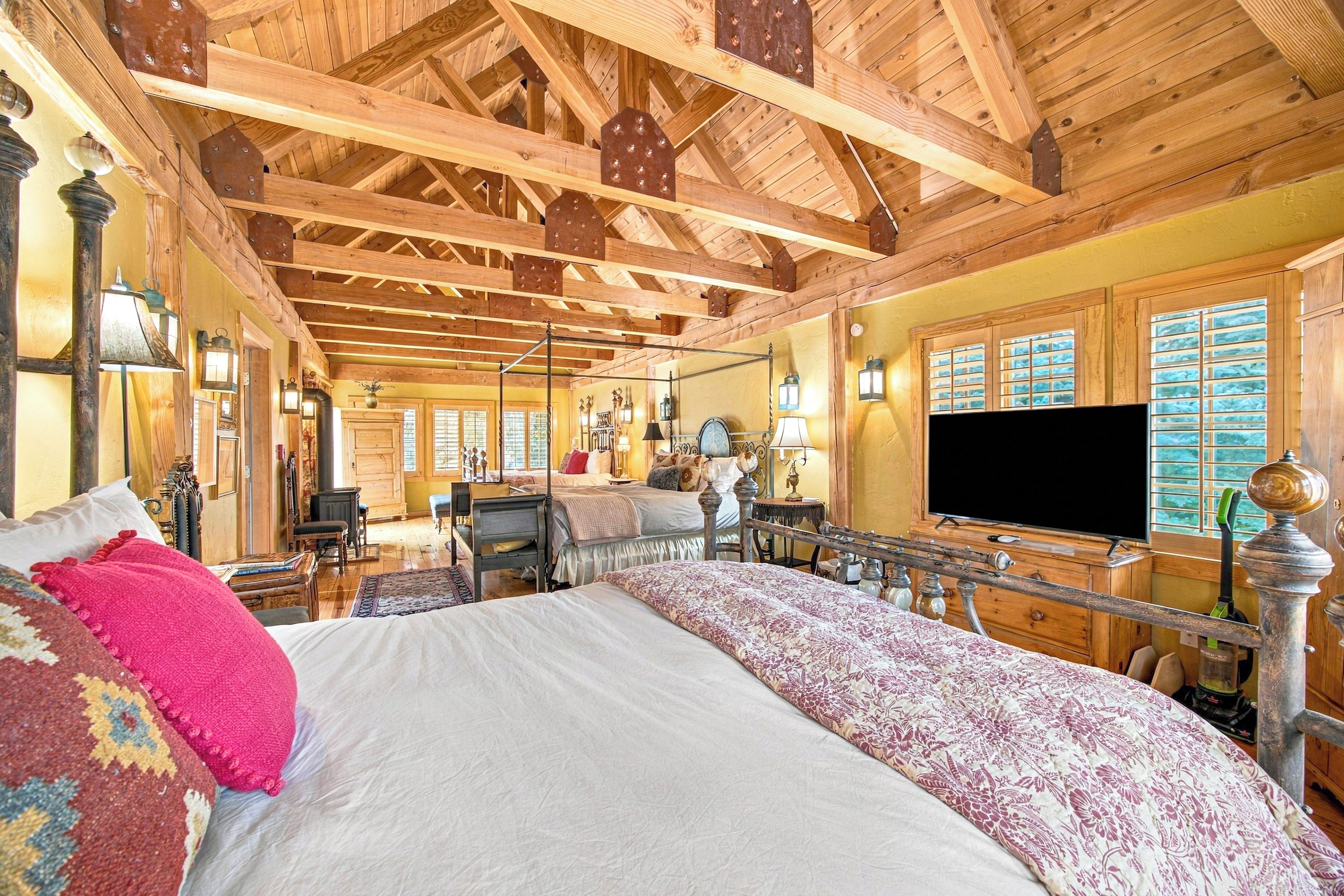 Un dormitorio amplio con una cama grande, vigas de madera en el techo, un televisor y vistas al exterior a través de las ventanas.
