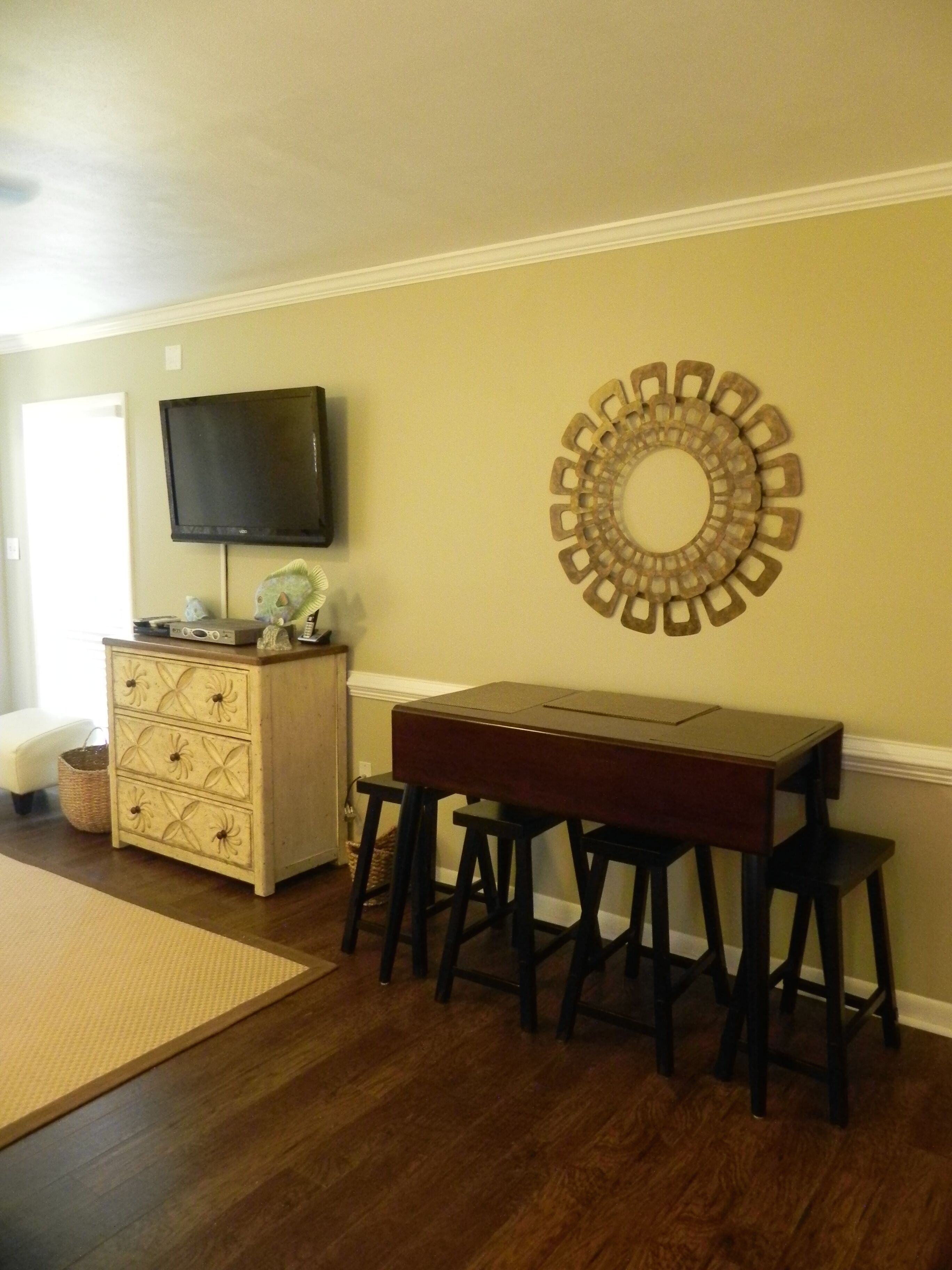 A room with a wooden table and four stools, a television on the wall, and a decorative circular wall piece.