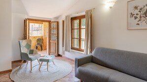 Un salon comprenant un canapé, un fauteuil, une petite table et un tapis. On y trouve une fenêtre avec des rideaux, un luminaire fixé au mur et un tableau encadré.