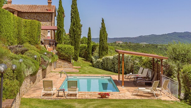 Une piscine avec des chaises longues, une pergola et un bâtiment en pierre surmonté d’un toit en tuiles.