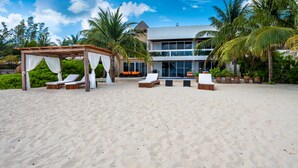 Un resort frente a la playa con un área de descanso cubierta, mobiliario exterior y un edificio moderno con grandes ventanales.