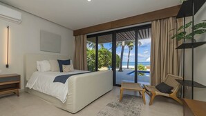 Un dormitorio moderno con cama, mesita de noche, una silla, una pequeña mesa y vistas al océano a través de amplios ventanales.