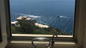 Blick durch ein Fenster auf eine Küstenlandschaft mit einer Bank im Vordergrund und einem Boot auf dem Wasser.
