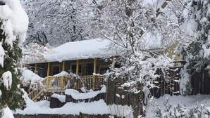 A snow-covered house with a stone wall foundation, wooden fence, and a yellow railing on the porch. - Fivespot Cabin, an art and nature lover's retreat (Pinehurst)