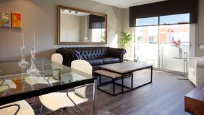 Un salon moderne avec une table à manger en verre, des chaises blanches, un canapé en cuir noir, un grand miroir et une vue sur la ville grâce à de grandes fenêtres.