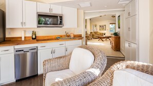 A modern kitchen with white cabinets, a stainless steel dishwasher, and a microwave. There is a wicker chair in the foreground and a dining area in the background. - Gulf Breeze (Naples)