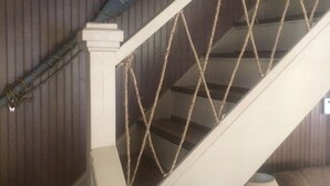 Un escalier avec une rampe blanche et une barrière faite de cordes, une lampe sur une table et une grille de ventilation au mur.