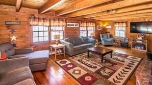 Interior acogedor de una cabaña de madera con chimenea, televisor y un amplio ventanal con cortinas.