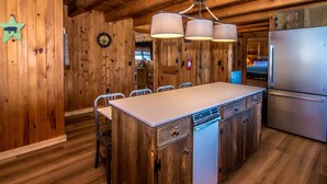 Un interior de madera con una zona de cocina que incluye una isla grande, sillas, un refrigerador y una lámpara en el techo.