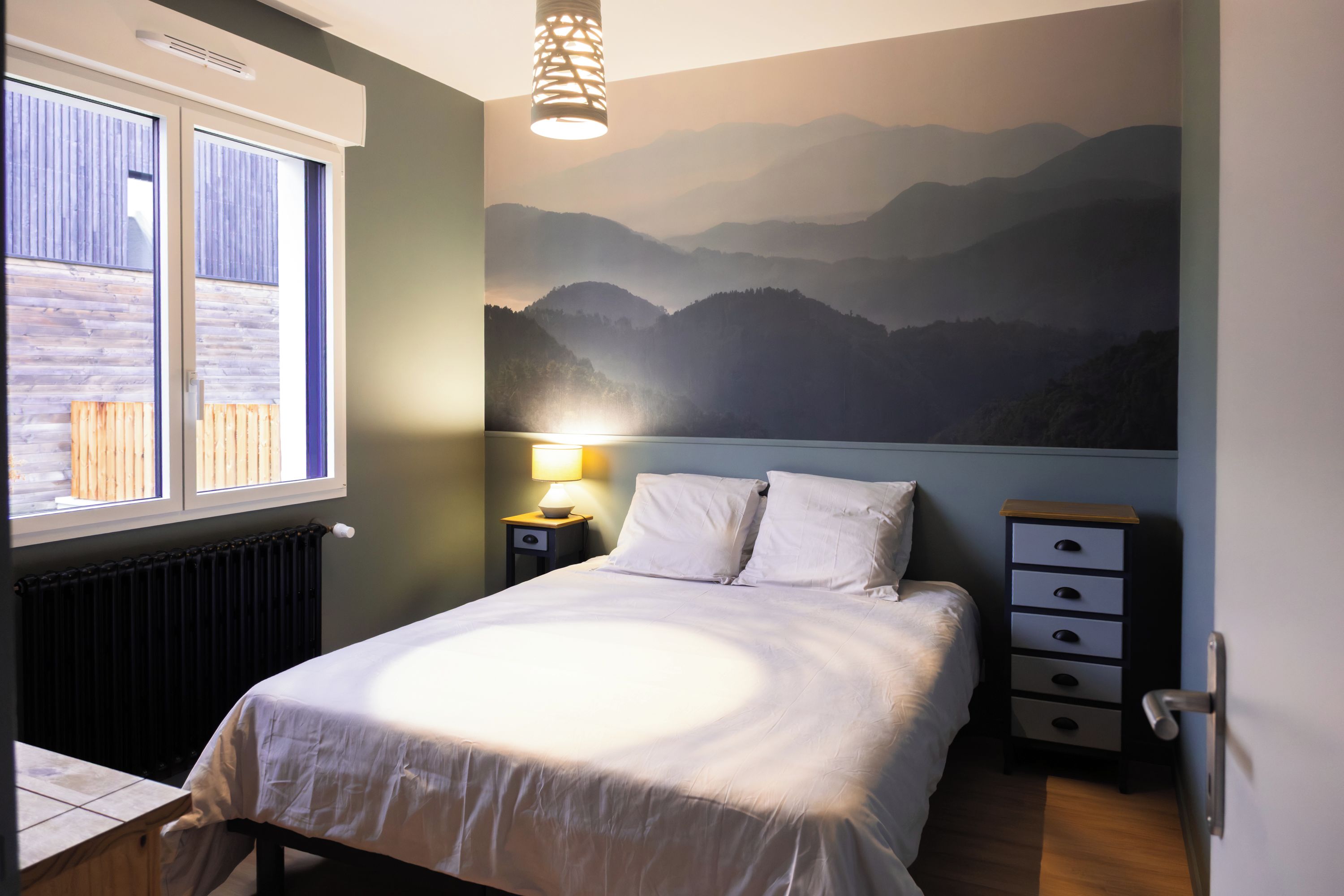 A bedroom with a bed, a nightstand, a lamp, a window, and a wall mural of mountains.