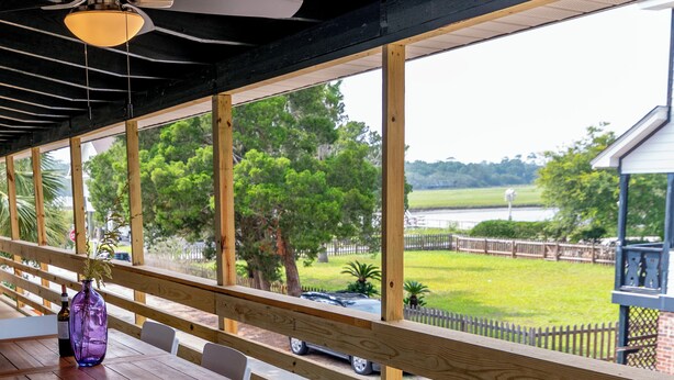 An outdoor seating area with wooden tables and chairs, a purple vase, and a view of a grassy area with trees and a fence. - Classic Pawleys. Just Renovated. Long, breezy porches. Steps to beach and creek. (Pawleys Island)