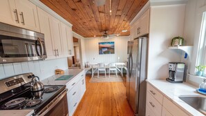 A modern kitchen with wooden flooring, white cabinetry, stainless steel appliances, and a dining area with a view. - Classic Pawleys. Just Renovated. Long, breezy porches. Steps to beach and creek. (Pawleys Island)
