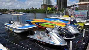 Marina con barcos, motos acuáticas y un edificio con letrero de alquiler de kayaks y tablas de paddle surf.