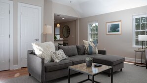 A living room with a gray sofa, a glass coffee table, and a patterned rug. There is a white door, a lamp, and a framed picture on the wall.