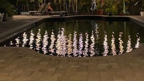 Un espace piscine extérieur éclairé la nuit par des guirlandes lumineuses, agrémenté de palmiers et offrant une vue sur un ciel dégagé.