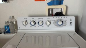 Une machine à laver à chargement frontal avec un panneau de commande, des bouteilles d’eau et une serviette bleue accrochée au mur.