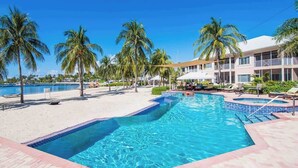 Une piscine dont le bord est revêtu de carrelage, entourée de palmiers et de chaises longues, avec un b âtiment visible au loin.