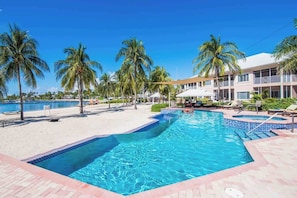 A swimming pool with a tiled edge, surrounded by palm trees and lounge chairs, with a building in the background. - Waterfront Korner Kai 2BR -- Pool, Jacuzzi & Beachfront Lanai (Cayman Kai)