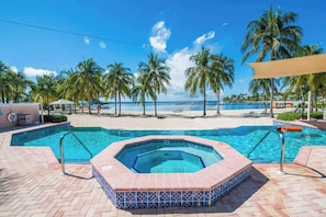 A swimming pool with a hot tub, surrounded by palm trees and a beach view.