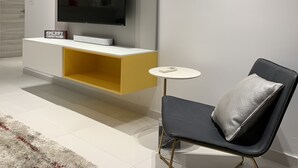 Een moderne woonkamer met een flatscreen televisie bevestigd aan een witte console, een klein rond tafeltje en een zwarte stoel met een zilveren kussen.