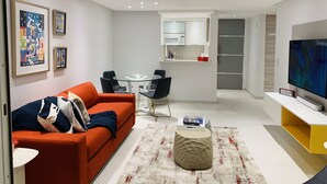 Een moderne woonkamer met een bank, salontafel en een televisie. Er is een eethoek met stoelen en een tafel. De keuken is zichtbaar, met een magnetron en kasten.
